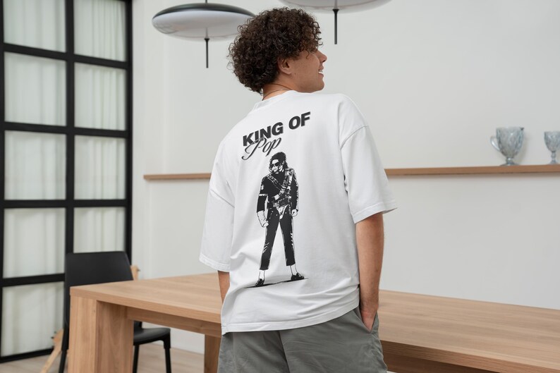 May include: White t-shirt with a black graphic of a person and the text "KING OF POP". The shirt is a short-sleeved, oversized style. The person in the image is wearing gray shorts.