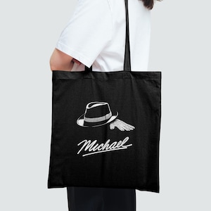 May include: Black tote bag featuring a white illustration of a fedora hat and a single glove. The word "Michael" is written in a cursive font below the graphic. The bag has black handles and is carried by a person.