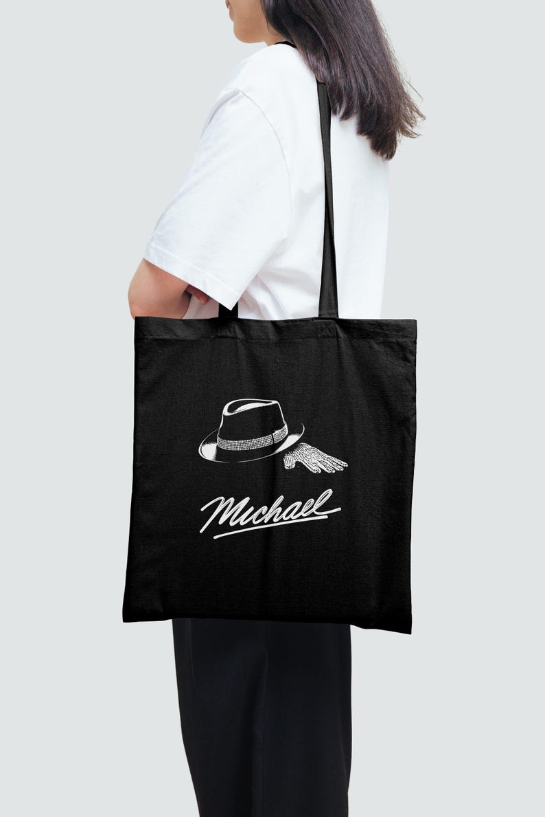 May include: Black tote bag featuring a white illustration of a fedora hat and a single glove. The word "Michael" is written in a cursive font below the graphic. The bag has black handles and is carried by a person.