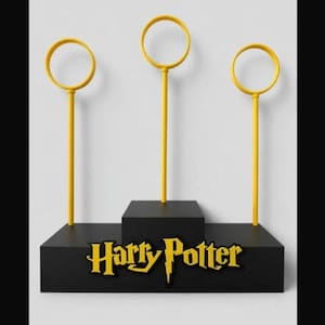 May include: A black display stand featuring three yellow Quidditch hoops. The stand has a tiered design and the words "Harry Potter" in gold lettering. The hoops are circular and mounted on slender poles.