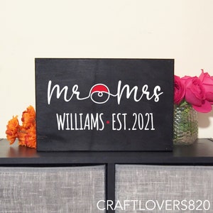 May include: Black wooden sign with white text that reads "mr & mrs Williams - est. 2021". A red and white Pokeball graphic is between the words "mr" and "mrs".