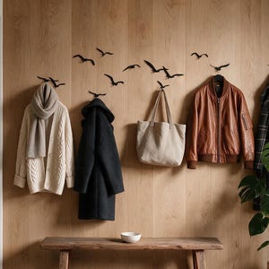 Op de afbeelding: Een set zwarte vogelvormige wandhaken gemonteerd op een wand met licht houten panelen. De haken dragen een beige kabelgebreid vest met bijpassende sjaal, een zwarte jas, een beige canvas tas, een bruine leren bomberjack en een geruite jas.
