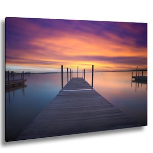 May include: A wooden pier extends into a calm body of water under a vibrant sunset sky. The horizon features orange and purple clouds reflecting on the smooth surface, with additional wooden dock structures visible on the left and right sides.