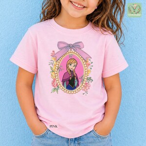 A young girl wearing a pink t-shirt with a picture of a girl in a pink dress and a bow on a blue background.