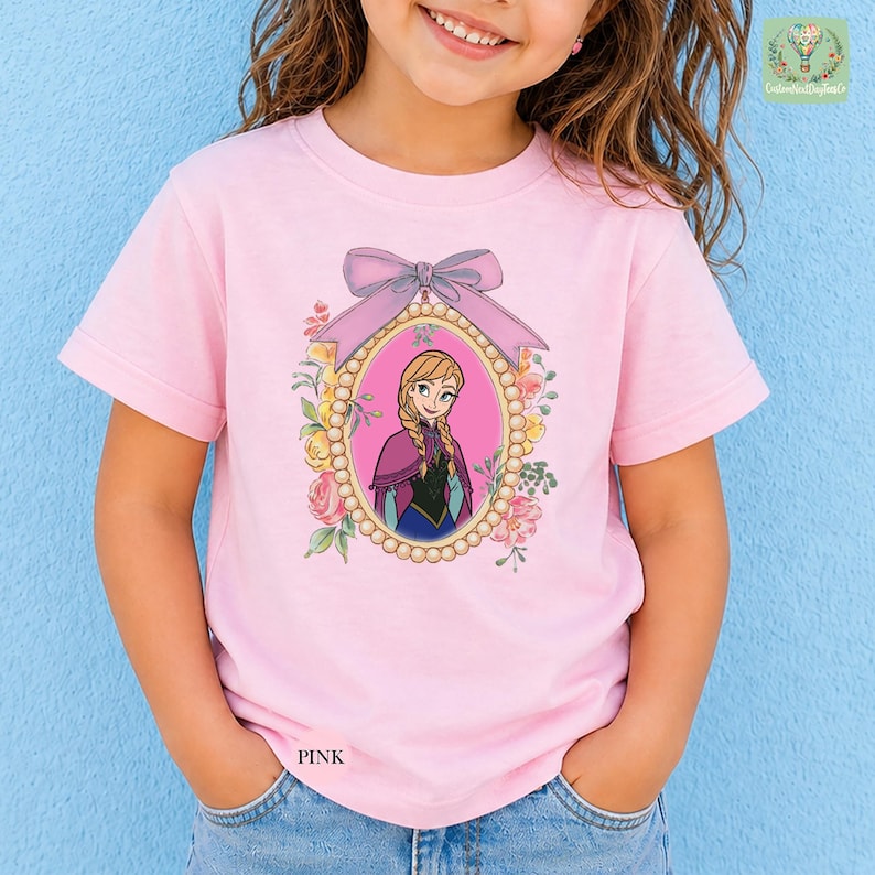 A young girl wearing a pink t-shirt with a picture of a girl in a pink dress and a bow on a blue background.