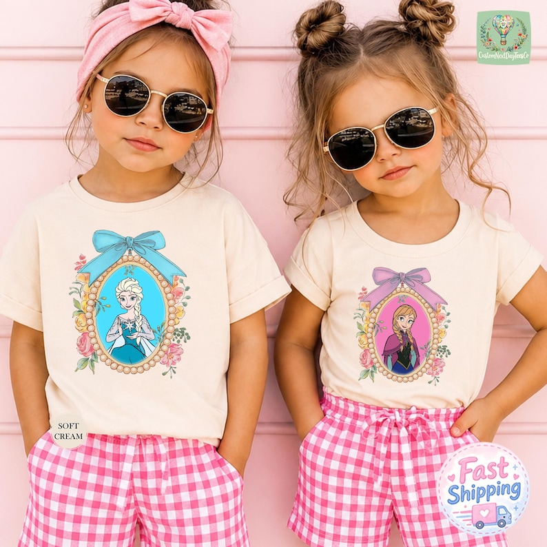 Two young girls wearing matching t-shirts with illustrations of characters from the Disney movie Frozen, standing against a pink background.