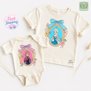 two white t-shirts with illustrations of two female characters from the Disney movie Frozen, one larger and one smaller. The larger t-shirt has a blue bow around the neckline, while the smaller t-shirt has a pink bow. The t-shirts are displayed on a white background, and there are some dried flowers and a shipping label visible in the top left corner.