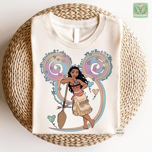 A white t-shirt with a colorful illustration of a young girl with dark hair and a pink dress, surrounded by whimsical elements such as flowers and a Mickey Mouse-like character.