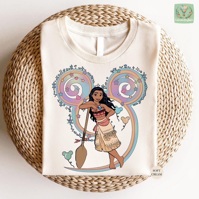 A white t-shirt with a colorful illustration of a young girl with dark hair and a pink dress, surrounded by whimsical elements such as flowers and a Mickey Mouse-like character.