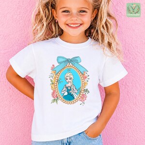 A young girl with blonde hair is smiling and posing in front of a pink wall. She is wearing a white t-shirt with a graphic of a character from the Disney movie Frozen.