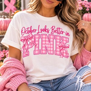 Op de afbeelding: Een wit T-shirt met korte mouwen en de roze tekst October Looks Better in PINK. Het woord PINK is geschreven in een groot bloklettertype met een roze en wit ruitpatroon, versierd met twee roze lintillustraties op de letters.