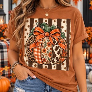 Könnte beinhalten: Ein rostfarbenes Kurzarm-T-Shirt mit einer großen Kürbisgrafik. Der Kürbis ist mit Abschnitten aus Leopardenmuster, orangefarbenem Karomuster, Blumenmustern und Kuhflecken verziert und wird von einer großen orangefarbenen Schleife gekrönt.