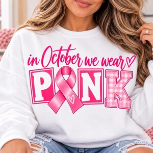 Op de afbeelding: Een wit sweatshirt met lange mouwen en de roze tekst "in October we wear" boven het woord "PINK" in grote blokletters. De letter "I" is vervangen door een roze lint voor borstkankerbewustzijn met een klein wit hartje.