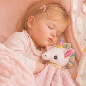 May include: A plush unicorn security blanket with a rainbow horn and a pink textured fabric body, alongside a cream-colored plush rabbit security blanket with a yellow neck ribbon, both featuring soft, dimpled fabric textures for tactile comfort.