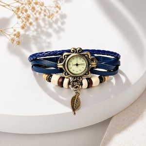 Puede incluir: Una pulsera envolvente de cuero azul con una esfera de reloj de estilo vintage. El reloj tiene una esfera color crema con números y manecillas negras. La pulsera también presenta cuentas de madera y un dije de hoja decorativo.