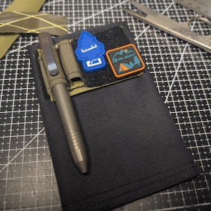 Pode incluir: Carteira de nylon preta com caneta, dois patches e um porta-canetas. A caneta é cinza metálico. Um patch é azul com um desenho de árvore branca, e o outro é laranja e azul com um desenho de tenda. A carteira está sobre uma base de corte preta.