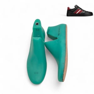 Puede incluir: Dos hormas de zapato de color verde azulado, talla 43, se muestran sobre un fondo blanco. Las hormas están hechas de un material sólido y están diseñadas para dar forma al calzado. Una zapatilla negra con rayas rojas y blancas se muestra en la esquina superior derecha.