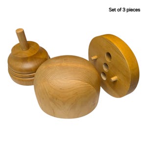 May include: A set of 3 pieces of light brown wooden objects. One piece is a rounded dome, another is a circular base with three holes and three small pegs, and the third is a rounded form with a vertical wooden handle protruding from the top.