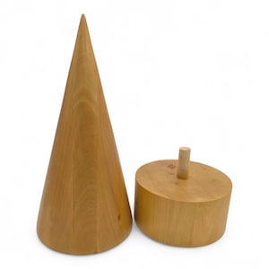 Puede incluir: Dos objetos de madera sobre un fondo blanco. Uno es un cono alto de color marrón claro. El otro es una forma cilíndrica corta con una pequeña clavija de madera en la parte superior. Los objetos son de madera natural.