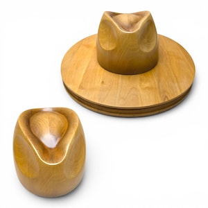 May include: A light brown wooden hat block with a smooth, polished finish. The set includes a circular base with a crown-shaped top piece and a separate, smaller, rounded crown-shaped block, both featuring a distinct, molded indentation.