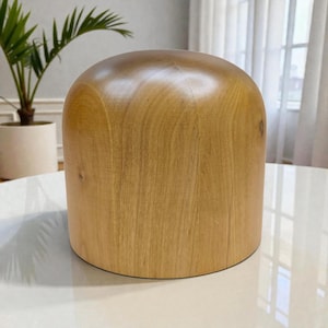 May include: A light brown wooden hat block, cylindrical in shape with a rounded top. The hat block is made of solid wood and has a smooth, polished finish. It is designed for shaping and blocking hats, a millinery tool.