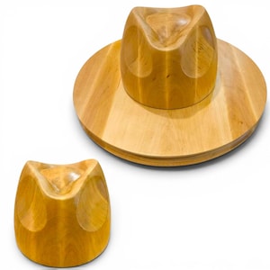 May include: A light brown wooden hat block with a smooth, polished finish. The object features a rounded crown with a deep, concave indentation at the top and a wide, circular brim base. A smaller, matching wooden form sits separately nearby.