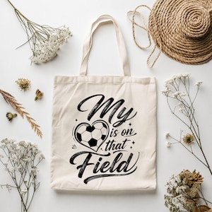 May include: A cream-colored canvas tote bag featuring a black graphic print. The design includes a soccer ball shaped like a heart and the text "My is on that Field" written in a stylized script font, centered on the front of the fabric bag.