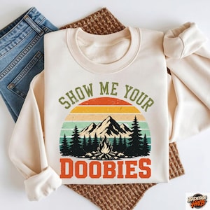 May include: Cream-colored sweatshirt with a graphic design featuring a mountain range, pine trees, and a campfire set against a sunset. The text "SHOW ME YOUR DOOBIES" is printed in green and orange. The sweatshirt is folded on top of blue jeans.