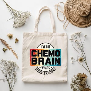 May include: A cream-coloured canvas tote bag featuring a central graphic with the text "I'VE GOT CHEMO BRAIN WHAT'S YOUR EXCUSE?" in bold black letters. The text is set against a rectangular background with a colourful, distressed gradient.