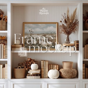 May include: A rectangular gold-toned picture frame containing a landscape print of a field with white pumpkins, displayed on a white shelf. The shelf features stacks of books, woven baskets, dried floral arrangements, and white pumpkins.