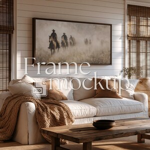May include: A framed artwork depicting cowboys on horseback in a misty landscape hangs above a white sofa with brown throw pillows and a knitted blanket. A wooden coffee table and a decorative vase are in front of the sofa. The text "Frame Mockup" is overlaid on the image.