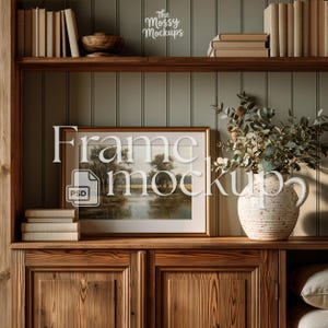 May include: A framed landscape painting with a gold frame is displayed on a wooden cabinet. A white ceramic vase with greenery sits to the right of the frame. The text "Frame Mockup" is overlaid on the image. The cabinet is part of a built-in bookshelf.