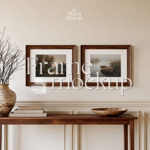 May include: Two horizontal rectangular dark wood frames with white matting, each containing a landscape print of a misty river scene with a small wooden boat. The frames are mounted on a beige wall above a wooden console table with a vase.