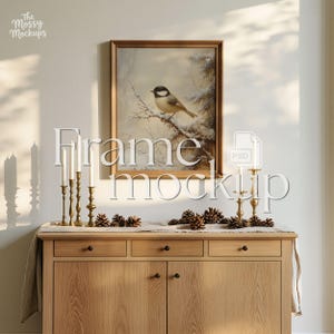 May include: A framed wall art print featuring a black-capped chickadee perched on a snowy branch, displayed above a light wood sideboard. The scene includes brass candlesticks with white candles and scattered pinecones on a linen runner.