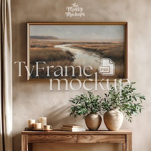 May include: A framed landscape painting with a gold frame hangs on a beige wall. The painting depicts a marsh scene with a river. Below, a wooden table displays candles, books, and vases with greenery. The text "TvFrame mockup" is overlaid on the image.