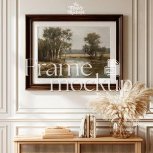 May include: A framed landscape painting with a dark brown wooden frame hangs above a wooden cabinet. The painting depicts trees and a body of water. A vase of dried pampas grass and a stack of books are on the cabinet. The text "Frame mockup" is overlaid on the image.