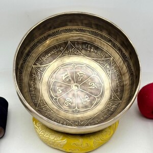 May include: A round, metallic singing bowl with a brass-toned finish. The interior features an etched lotus flower design with eight petals, each containing a symbol, surrounded by a geometric star pattern and intricate Sanskrit-style text.