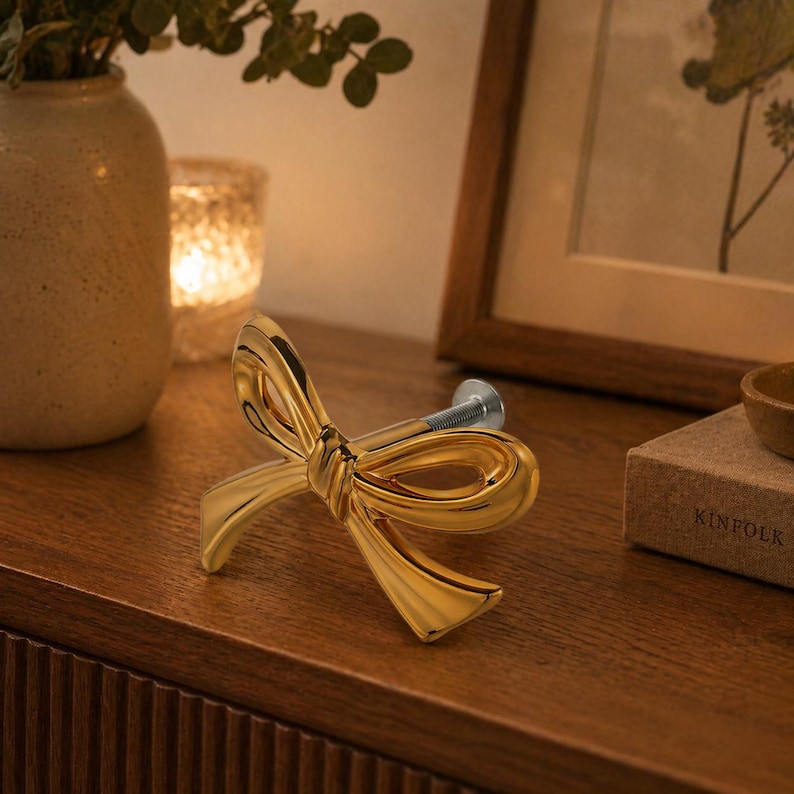 May include: A gold-coloured bow-shaped cabinet pull with a silver screw. The decorative hardware is made of metal and is designed to be installed on a drawer or cabinet door. The bow has a glossy finish.