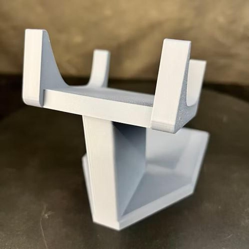 May include: A light gray 3D printed stand featuring a flat rectangular top platform with four vertical U-shaped supports at the corners, connected to a solid, angular, tapered base that provides stability for the elevated upper section.