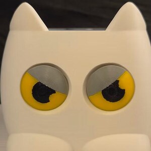 May include: A white, cat-shaped container with two large, circular eyes. Each eye features a grey upper half, a yellow lower half, and a black center pupil. The object has two pointed ears at the top and two rounded paws at the bottom base.