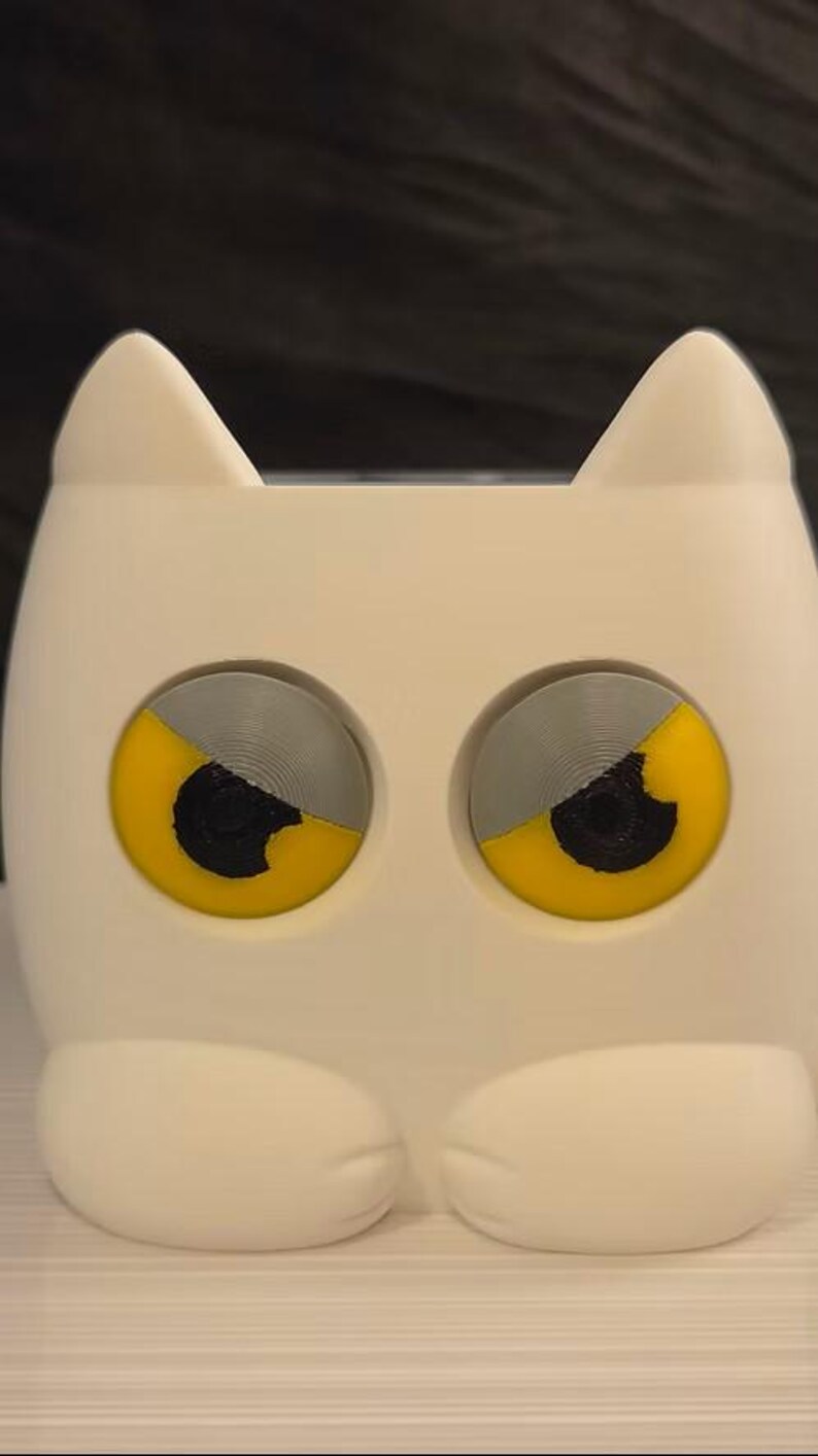 May include: A white, cat-shaped container with two large, circular eyes. Each eye features a grey upper half, a yellow lower half, and a black center pupil. The object has two pointed ears at the top and two rounded paws at the bottom base.