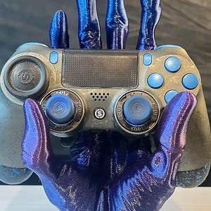 May include: A dark blue, glittery, hand-shaped stand holding a gray and blue video game controller. The controller features a central touchpad, blue action buttons, and two analog sticks with the text SCUF printed on the circular base of each stick.