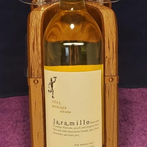 May include: A bottle of 2023 Jaramillo Moscato red wine, 750 ml, with a label describing sweet notes of orange blossom, peach, and tropical fruit. The bottle is held in a wooden wine glass holder.