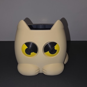 May include: A cream-colored cat-shaped planter with large, round eyes featuring black pupils and yellow irises. The container has two pointed ears at the top and two rounded paws at the base. The interior of the planter is dark gray and hollow.