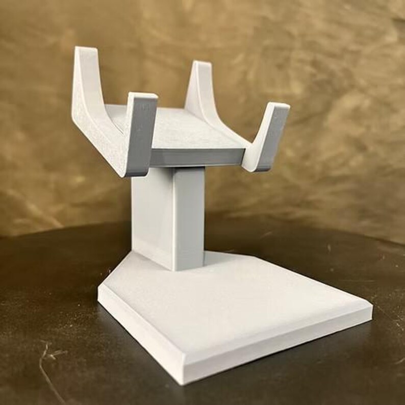 May include: A light gray display stand featuring a wide, flat pentagonal base, a central vertical support pillar, and a top cradle section with two curved, upward-extending arms designed to hold an object securely in a stable, elevated position.