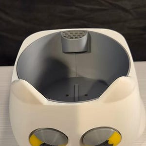 May include: A white, rounded container with two pointed ears at the top and two large, circular eyes featuring yellow irises and grey pupils. The interior is grey with a small, perforated semi-circular tab at the top edge and a flat base.