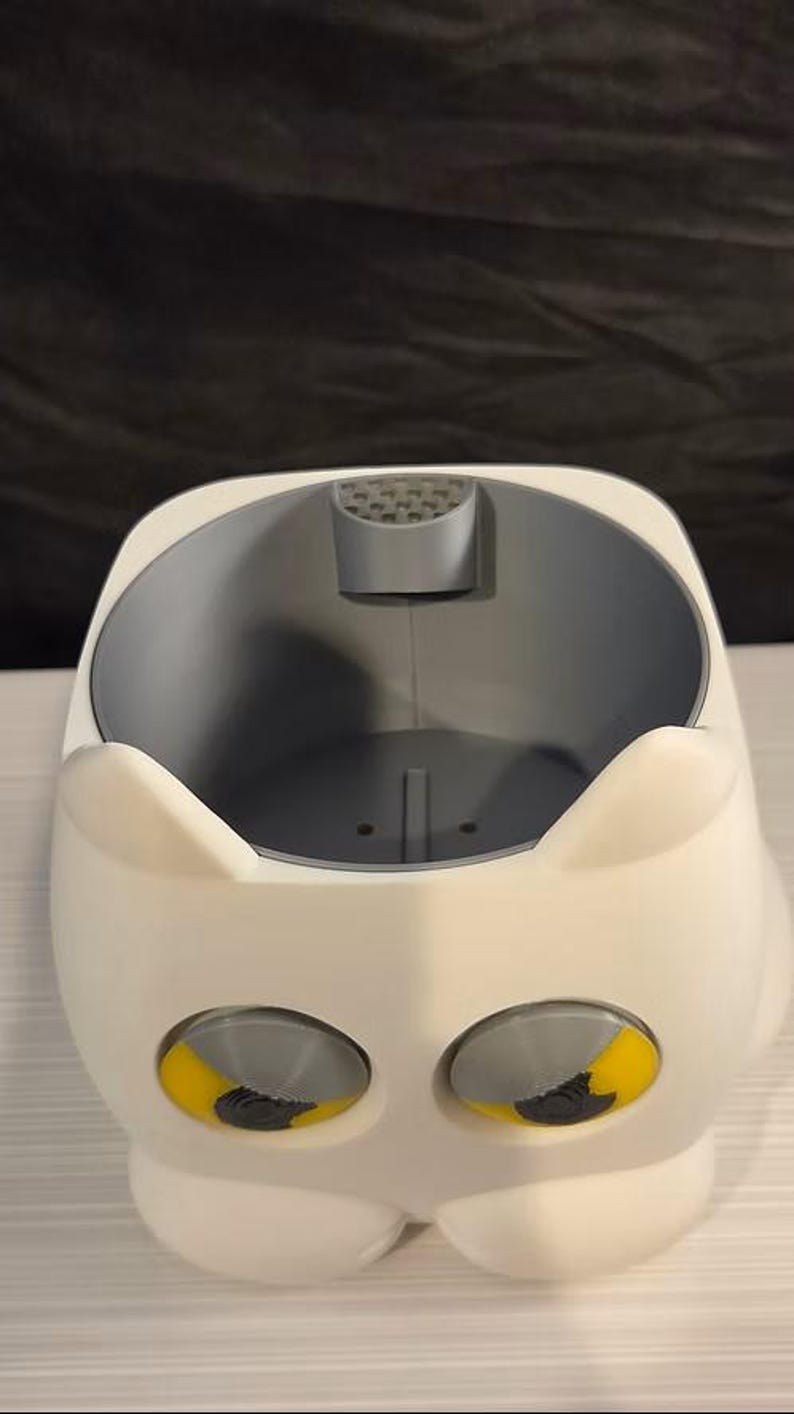 May include: A white, rounded container with two pointed ears at the top and two large, circular eyes featuring yellow irises and grey pupils. The interior is grey with a small, perforated semi-circular tab at the top edge and a flat base.
