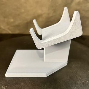 May include: A white 3D-printed display stand featuring a flat rectangular base and an angled support arm with two upward-curving prongs designed to hold an object. The surface of the stand has a visible textured finish from the printing process.