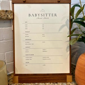 May include: A wooden clipboard with gold-toned metal accents holding a cream-colored paper titled BABYSITTER cheat sheet. The form includes sections for kids, tonight, important, contact, good to know, and house information with blank lines.