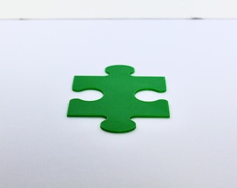 May include: A single, solid green puzzle piece with four interlocking tabs and notches. The piece is flat and features a smooth, matte surface texture, positioned centrally against a plain, light-colored, neutral background.